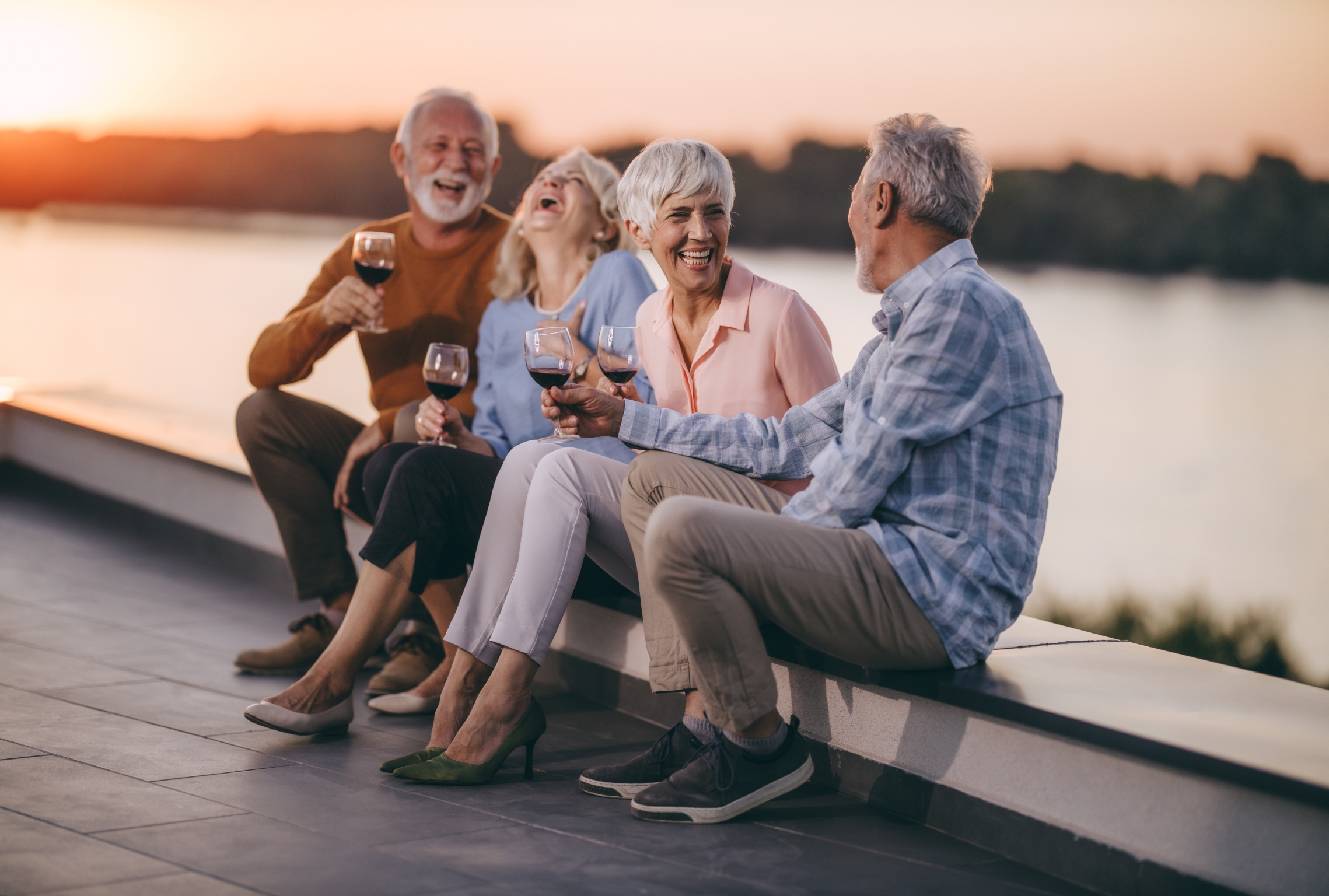 two senior coupls enjoying wine on a ledge near a lake