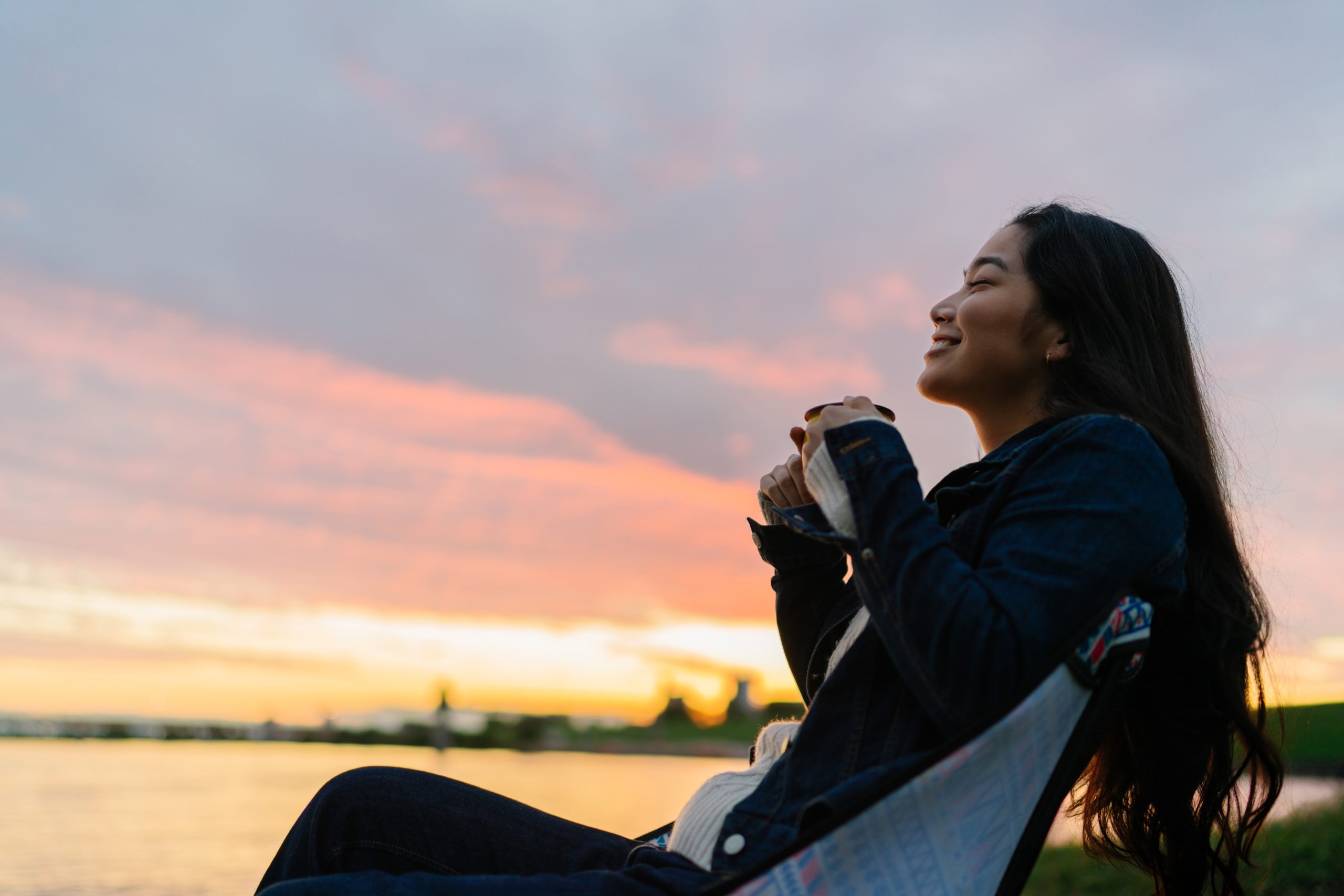 A young woman is enjoying sitting near a lake and drinking a hot drink in nature during sunset.