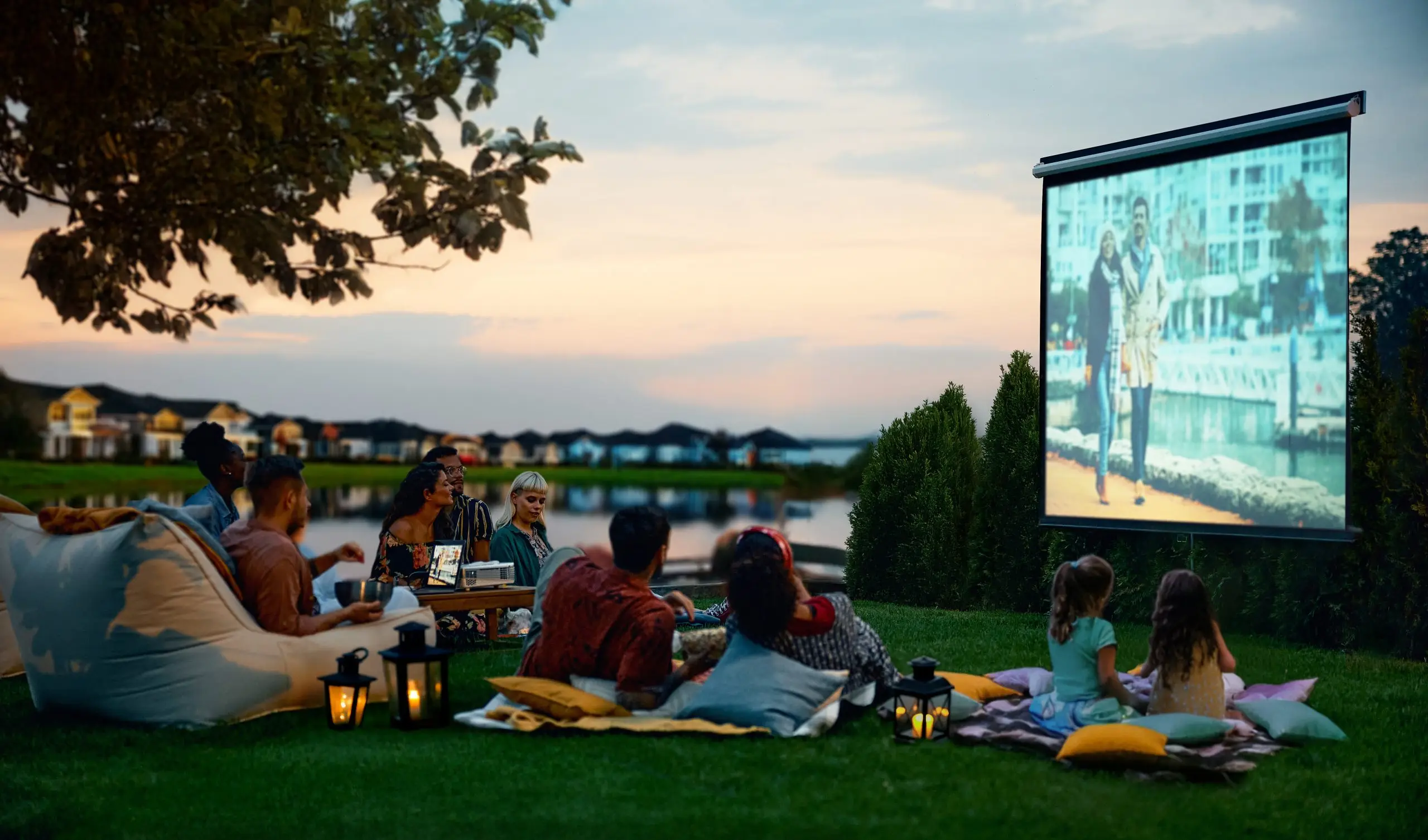 adults and children sitting on pillows and blankets by a lake watching an outdoor movie on a screen