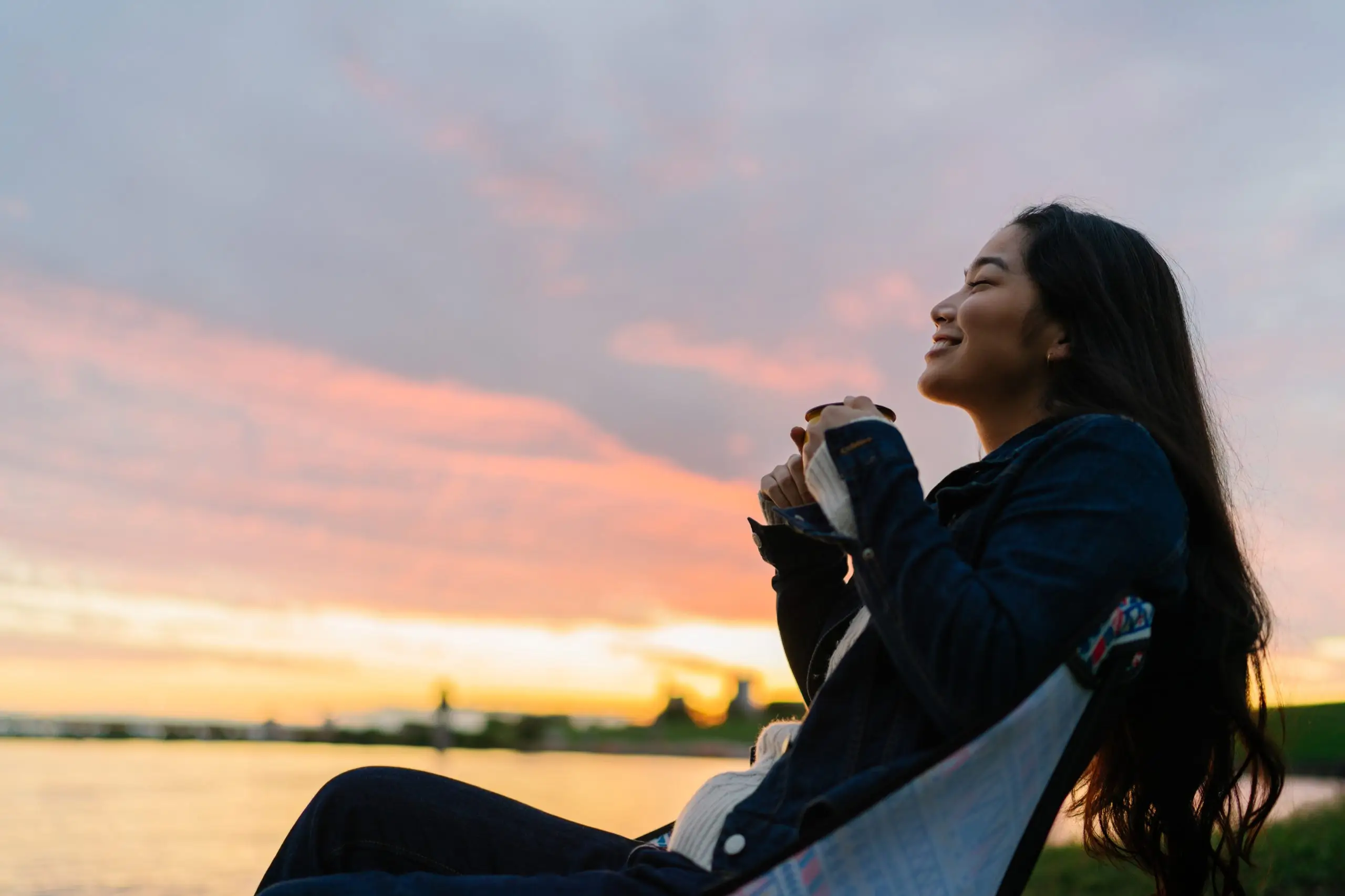 A young woman is enjoying sitting near a lake and drinking a hot drink in nature during sunset.