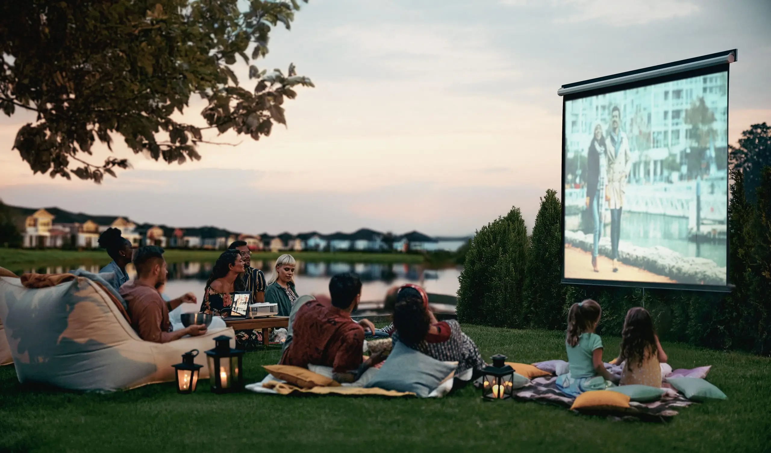 adults and children sitting on pillows and blankets by a lake watching an outdoor movie on a screen