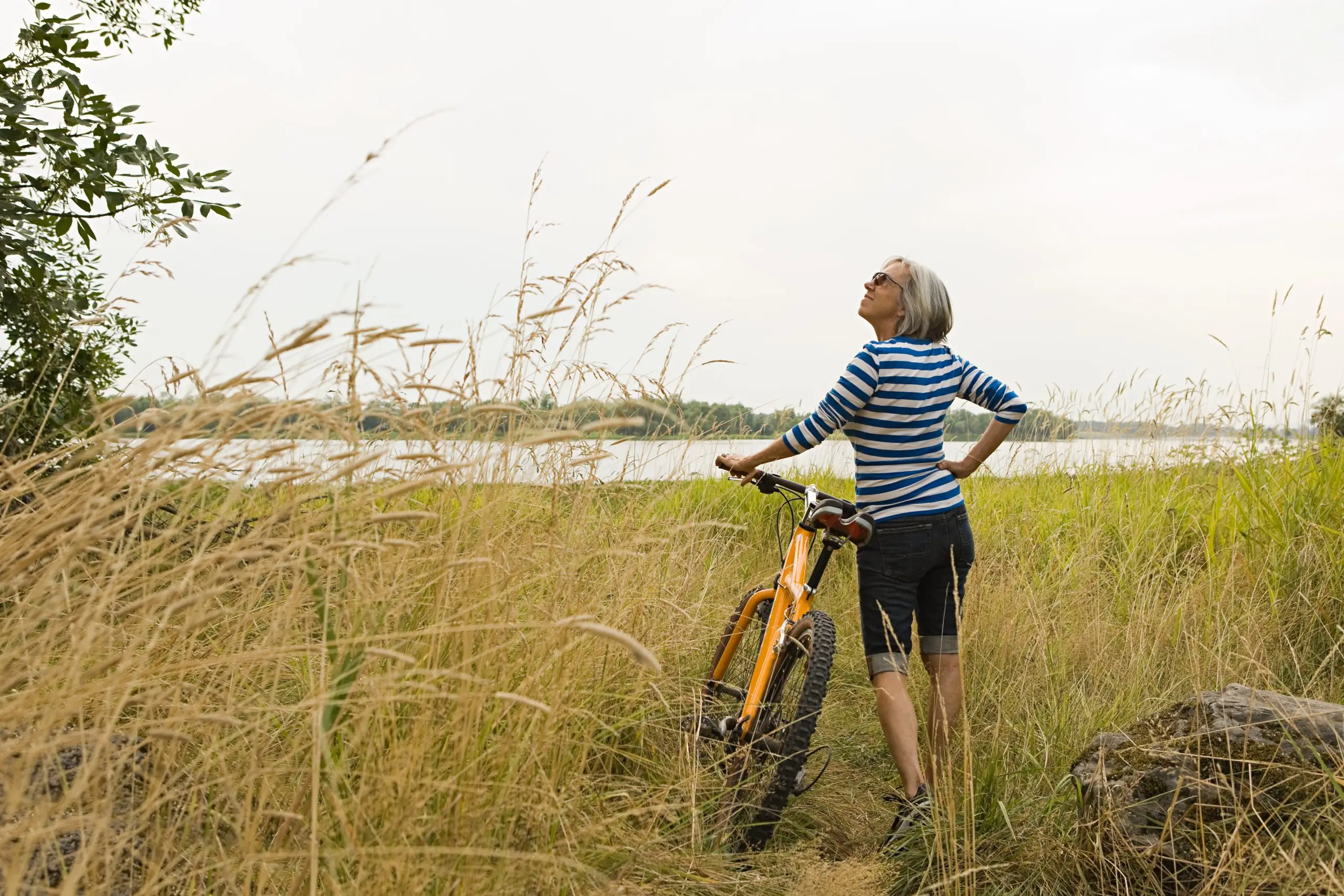 woman stopping to look up while walking with her bike in foliage next to a lake