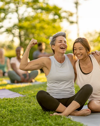 woman flexing muscle on a yoga mat outside with friend